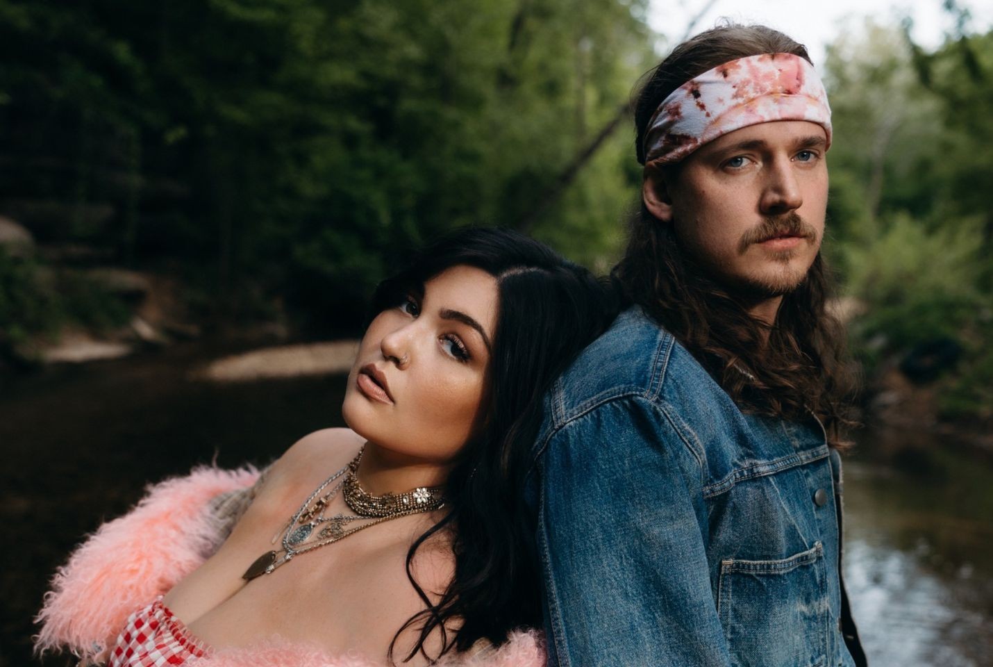 A man and woman stood back to back in front of a river. the woman is wearing a pink fluffy coat around her shoulders and lots of silver necklaces. The man has on a blue denim jacket and pink and white tie dye bandana around his head